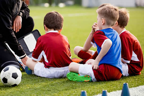 An image of a group of young players sitting on the ground listening to their coach